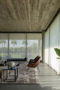 A minimalist living room with concrete ceiling and floor, featuring large windows, a brown armchair, a dark coffee table, and