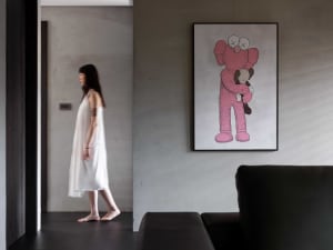 A woman wearing a white dress and walking past a wall with a large framed drawing of a pink teddy bear.