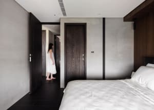 A minimalist bedroom with concrete walls, a white bed, and a person walking through the hallway.