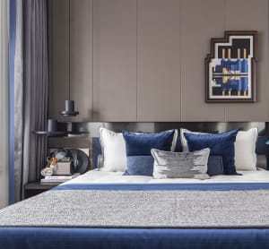 A modern bedroom featuring a bed with blue and white textiles, a nightstand with headphones, and a framed artwork on the wall