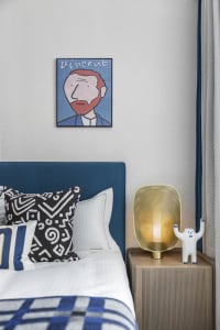 A modern bedroom featuring a blue upholstered headboard with patterned throw pillows and a golden lamp on a side table.