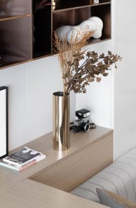 A metallic vase containing dried flowers sits on a wooden surface, with a camera and framed artwork nearby.