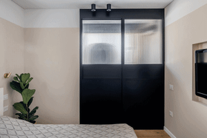 A minimalist bedroom featuring black steel and glass sliding doors with a wooden bed and green plant.