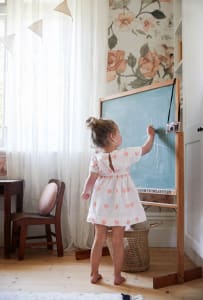 A young child stands at a wooden easel drawing on a chalkboard in a cozy, flower-patterned children's room.