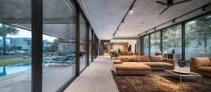 Modern living room with concrete walls and floor, featuring a brown leather sofa and a rug, with a pool area visible through 