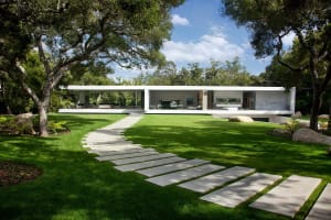 A modern minimalist house with white exterior, glass walls, concrete patio, and greenery