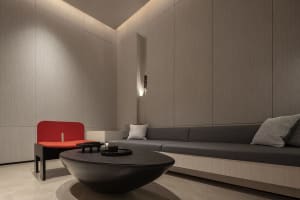 A minimalist living room with a curved concrete coffee table, a red accent chair, and a built-in gray sofa with patterned thr