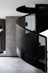Industrial-inspired spiral staircase with a perforated metal balustrade and black metal handrail, set against a minimalist ba
