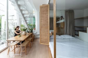 A modern minimalist bedroom featuring a large bed with white sheets, a wooden table with a vase and plants, and a large windo