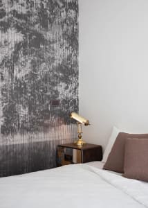 Minimalist hotel room featuring a textured wallpaper wall and a brass table lamp on a wooden bedside table.