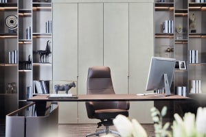 Modern office desk space featuring a wooden and leather chair, a desk with a computer monitor, and a bookshelf filled with bo