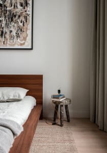 A minimalist bedroom featuring wooden furniture and a hanging light bulb on a stool, with a framed abstract painting above th