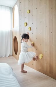 A toddler climbing on a wooden wall with yellow climbing holds in a modern children's playroom.