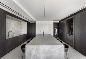 A modern residential kitchen with a long marble island centerpiece, dark wooden cabinetry, and sleek black chairs.