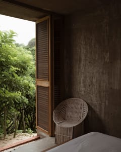 A modern bedroom featuring a concrete wall and a macrame chair, with a view of lush greenery outside through an open window.