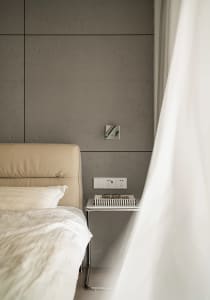 A minimalist bedroom featuring a bed with a beige leather headboard and a nightstand with a book, glasses, and power outlets 