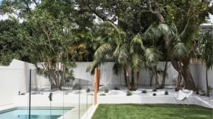 Modern poolside area with white stonework, glass fencing, and lush greenery including palm trees.