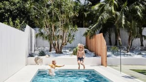 A child playing in a modern backyard pool surrounded by palm trees and a white fence.