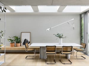 A minimalist dining room with a white table, cane chairs, and a white wall with skylights.