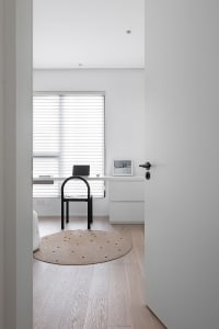 A minimalist home office with a wooden desk and black chair, featuring a round rug and a window with blinds.