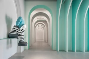 Blue sculpture with a striped black and white body sitting in a minimalist corridor with white and aqua walls.