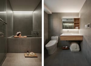 A modern bathroom featuring a gray tiled shower area with a person lying in a tub, a white toilet, and a vanity with a wooden