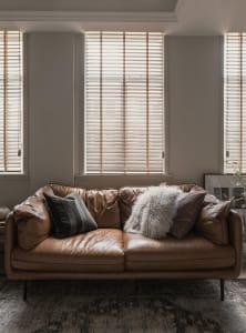 A modern leather sofa adorned with throw pillows and placed in front of a window with wood blinds.