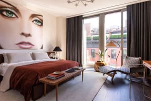 Modern bedroom with a large wall mural of a woman's face, wooden bed frame, fabric bedding, metal floor lamp, and wooden benc