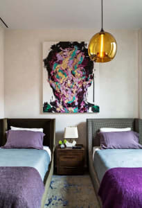 A modern bedroom with two beds, purple linens, a wooden bedside table with a lamp and small plant, and a large abstract paint