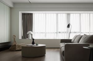 A modern living room with a light wood floor and a large floor-to-ceiling window with sheer curtains.