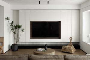 Modern, minimalist living room with white walls, a black marble shelf, a round wooden coffee table, and a flat-screen TV embe