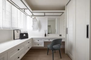 A minimalist white clothing closet with a hanging rod, drawers, and a desk area with a mirror and chair.