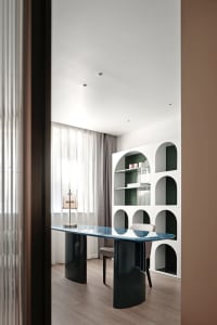 Modern office space featuring a sleek glass and metal table, arched shelving unit, and a window with sheer curtains.