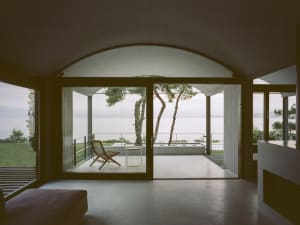 A modern interior space with glass and concrete elements, featuring a lounge chair and view of a peaceful outdoor area.