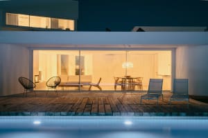 A modern vacation home at night with a wooden deck, glass walls, and a swimming pool.