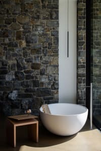 A minimalist bathroom with a stone wall, white bathtub, wooden stool, and glass door.