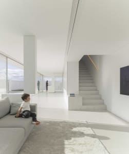 A minimalist living room with white walls, concrete floors, and a large window allowing natural light to fill the space.