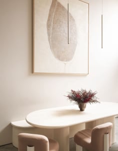 A minimalist dining room with a wooden table and chairs, adorned with a flower arrangement and a large abstract painting.