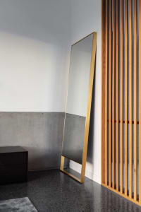 A modern floor mirror with a gold metallic frame leaning against a wall in a minimalist interior setting.