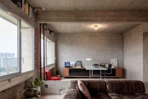 A modern industrial living room with a concrete wall, a leather sofa, a red chair, and a desk with various items.