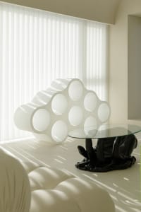 A minimalist space featuring a modern, glass coffee table with a hand-shaped base and a white, cloud-like structure with circ