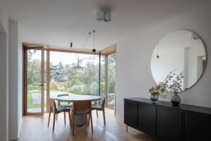 A modern minimalist dining area with a round table, chairs, and a large window overlooking a garden.