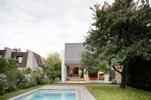 A modern residential backyard featuring a house with a pool and a large tree. The house has a minimalist design with wooden d
