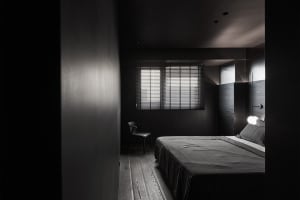 Minimalist bedroom with wood flooring and furniture, featuring a single window with blinds and a lit lightbulb.