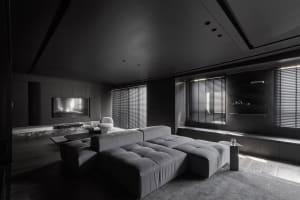 Minimalist living room interior with a upholstered grey couch, recessed lighting, and black window shutters.