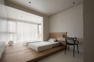 A minimalist bedroom featuring twin beds on a wooden platform with sheer curtains and a black chair against a white wall.