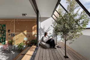 A woman sits on a cushioned chair reading a paper in a modern living space with wooden flooring and plants.