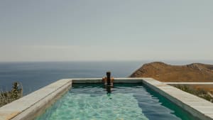 A woman sitting in a pool overlooking a scenic landscape with mountains and sea.