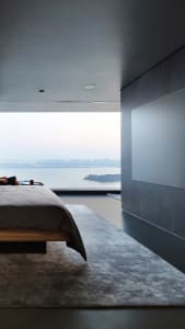 A minimalist bedroom featuring a concrete floor and a large window overlooking a cityscape.