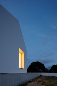 A modern white house with geometric concrete cuts, seen at dusk with illuminated windows.